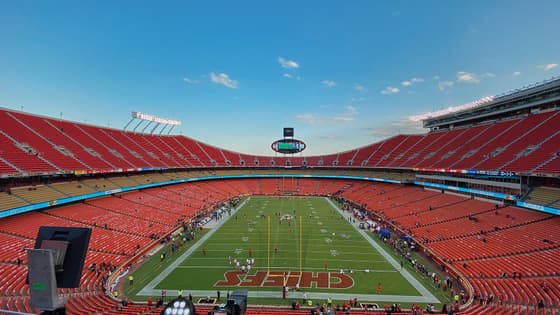 Vista panorámica del interior del Arrowhead Stadium de Kansas City antes del partido: gradas rojas casi vacías, el campo de pasto con la palabra CHIEFS pintada en la zona de anotación y cielo azul sobre el estadio.