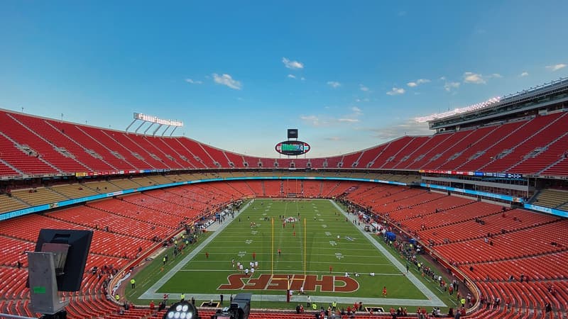 Vista panorámica del interior del Arrowhead Stadium de Kansas City antes del partido: gradas rojas casi vacías, el campo de pasto con la palabra CHIEFS pintada en la zona de anotación y cielo azul sobre el estadio.