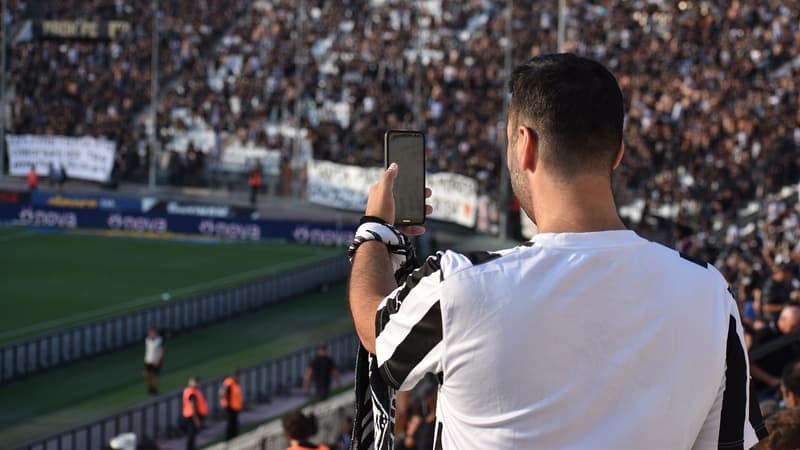 Un aficionado de espaldas sostiene su celular en la tribuna de un estadio de futbol lleno antes del partido.