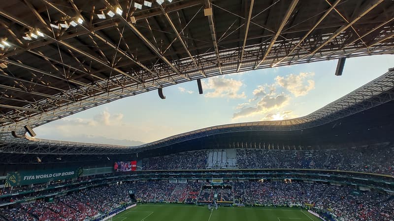 Interior del Estadio BBVA de Monterrey al atardecer, con las gradas llenas, la cancha iluminada y el techo ondulado visto desde la parte alta.
